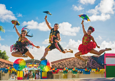 Imagem: Fotografia. Três pessoas, no ar, com a perna direita dobrada e o braço direito erguido, seguram um guarda-chuva pequeno e colorido.  Fim da imagem.