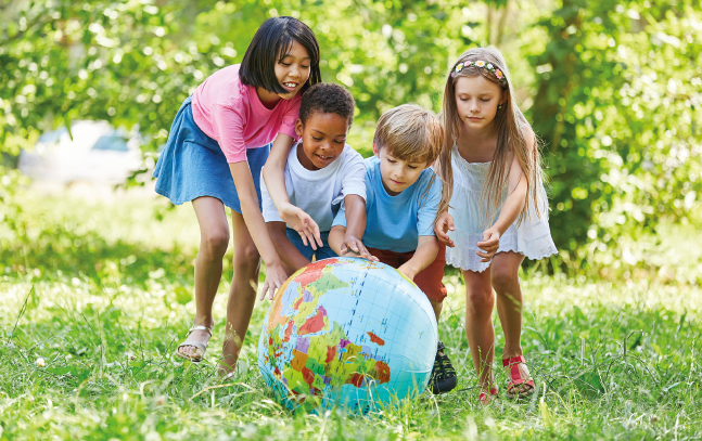Imagem: Fotografia. No centro, crianças abaixadas, com os braços esticados para frente, tocam um globo terrestre que está no chão. Ao fundo, árvores e grama. Fim da imagem.