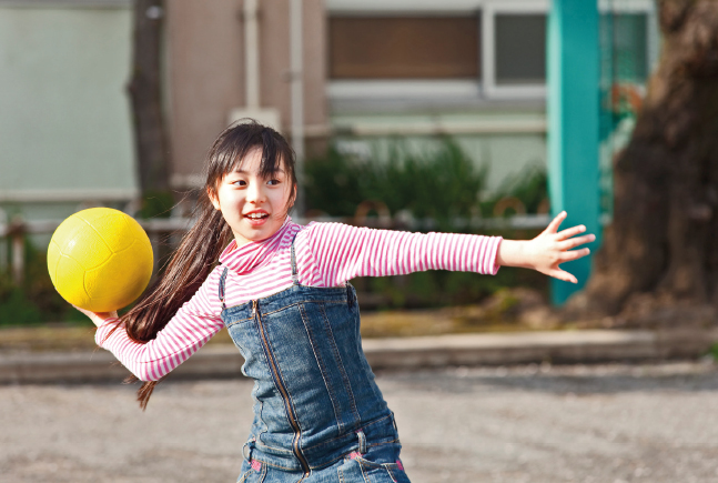 Imagem: Fotografia. Uma menina de cabelo longo com franja, veste camisa listrada rosa e macacão jeans, está com os braços abertos e segura uma bola amarela com a mão direita.  Fim da imagem.