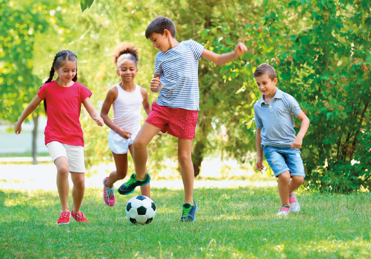Imagem: Fotografia. No centro, um menino vestindo camisa listrada branca e preta e bermuda vermelha, está com o pé direito levantado na direção de uma bola de futebol. Atrás dele, três crianças olham para a bola. O chão é de grama e ao fundo há árvores. Fim da imagem.