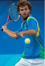 Imagem: Fotografia. Um homem, vestindo camiseta azul, está com o braço direito flexionado para trás, segura uma raquete com a mão direita e olha para uma bola verde.  Fim da imagem.