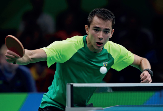 Imagem: Fotografia. Um homem, vestindo camiseta verde, está com os braços abertos, segura uma raquete pequena na mão direita e olha para uma bola branca. À sua frente, uma rede esticada.  Fim da imagem.