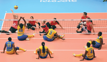 Imagem: Fotografia. Na frente, seis mulheres sentadas estão vestindo camiseta amarela e olhando para uma bola amarela no alto. No meio, uma rede esticada com bordas brancas. Ao fundo, outras seis mulheres estão sentadas, vestem camiseta vermelha e olham para a bola.  Fim da imagem.