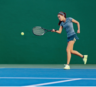 Imagem: Fotografia. Uma mulher está com o braço direito dobrado para frente e segura uma raquete na direção de uma bola verde.  Fim da imagem.