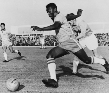 Imagem: Fotografia em preto e branco. Um homem, vestindo camiseta, bermuda e meia alta, está com o joelho direito dobrado, a perna esquerda esticada, os braços estendidos e olha para uma bola no chão. Ao redor dele, outros homens olham para a bola.  Fim da imagem.