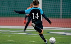Imagem: Fotografia. Uma pessoa está com os braços abertos, perna direita levantada e próxima de uma bola de futebol. Fim da imagem.