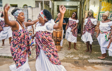 Imagem: Fotografia. No centro, duas mulheres, com as sobrancelhas levantadas e sorrindo, vestindo roupa branca e lenço florido na cintura, estão com os braços levantados e próximas uma da outra. Ao redor, mulheres com a mesma roupa.  Fim da imagem.