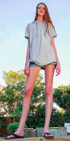 Imagem: Fotografia. Uma mulher com pernas compridas, cabelo longo, camiseta cinza e bermuda está olhando para baixo.   Fim da imagem.