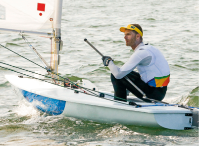 Imagem: Fotografia. Um homem com boné amarelo, blusa branca e calça preta está sentado em um barco a vela.   Fim da imagem.