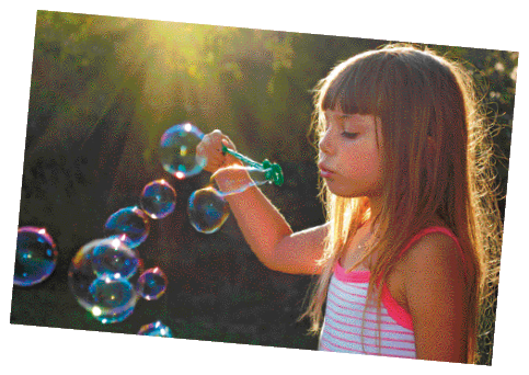 Imagem: Fotografia. Uma menina com cabelo comprido e regata está segurando um aro e assoprando. Na frente dela há várias bolhas de sabão.  Fim da imagem.