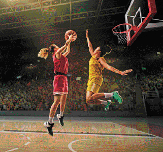 Fotografia. Uma mulher com uniforme vermelho está pulando e segurando uma bola de basquete. Na frente dela, uma mulher com uniforme amarelo está pulando com o braço para cima. Atrás delas há uma cesta e ao fundo, pessoas em uma arquibancada.