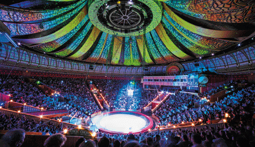 Fotografia. Interior de um picadeiro. No centro, o palco circular e em volta, várias pessoas em arquibancadas. No teto há luzes e faixas coloridas.