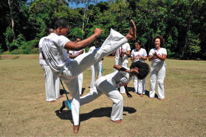 Fotografia. No centro, duas pessoas estão lutando capoeira. Atrás, pessoas batem palma. Ao fundo, árvores.