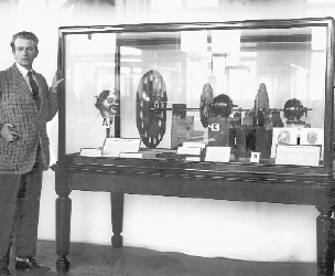 Imagem: Fotografia em preto e branco. Um homem com terno está ao lado de uma televisão grande e larga.   Fim da imagem.