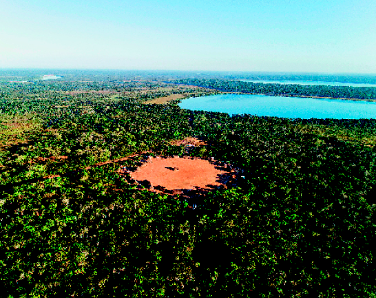 Imagem: Fotografia. Vista aérea de uma floresta. No centro há uma área com ocas em círculo. Ao fundo, um lago.  Fim da imagem.