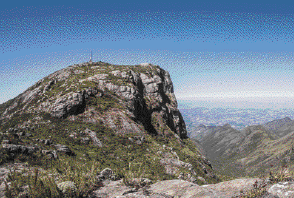 Imagem: Fotografia. Pico cinza com vegetação. Ao fundo, o céu azul.  Fim da imagem.