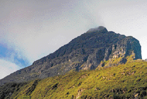 Imagem: Fotografia. Pico coberto de vegetação. Ao fundo, nuvens escuras.  Fim da imagem.