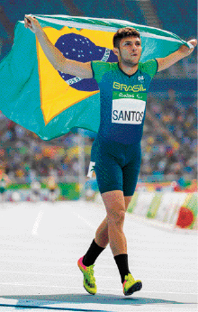 Imagem: Fotografia. Um homem com camiseta e bermuda verde-escuro está andando e segurando a bandeira do Brasil atrás das costas. Fim da imagem.