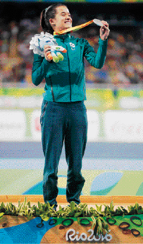 Imagem: Fotografia. Mulher com cabelo preso, casaco e calça verde-escuro está sorrindo sobre um pódio e segurando uma medalha, que está pendurada no seu pescoço. Fim da imagem.