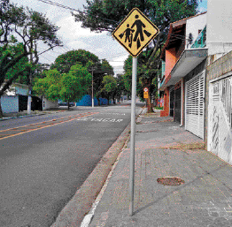 Imagem: Fotografia. Placa amarela com formato de losango. No centro, silhueta de um adulto e uma criança atravessando sobre a faixa de pedestres. Ao fundo, uma rua e casas.   Fim da imagem.