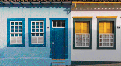 Imagem: Fotografia. Casas lado a lado com janelas e portas retangulares e coloridas.  Fim da imagem.