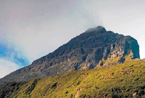 Imagem: Fotografia. Pico coberto de vegetação. Ao fundo, nuvens escuras.  Fim da imagem.