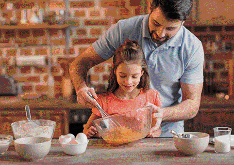 Imagem: Fotografia. Um homem está atrás de uma menina e eles estão segurando uma tigela e um batedor. Na frente deles há vários ingredientes sobre uma mesa. Fim da imagem.