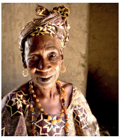 Imagem: Fotografia. Uma mulher negra com cabelos brancos e rugas, usando um turbante e um vestido estampado com corrente no pescoço. Fim da imagem.