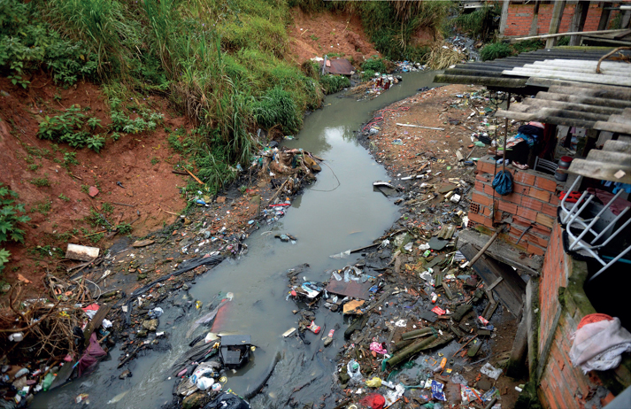 Imagem: Fotografia em Página dupla. À esquerda, margem de terra com lixo. No centro, um córrego com muito lixo dentro e nas margens. À direita, paredes de casas com telhado.   Fim da imagem.