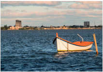 Imagem: Fotografia. Um barco pequeno em uma lagoa. Atrás, areia e silhuetas de dois prédios. Fim da imagem.