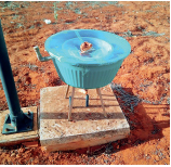 Imagem: Fotografia. Um balde azul, sobre um tripé de madeira, com uma tampa e uma válvula à esquerda.  Fim da imagem.