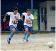 Imagem: Fotografia. Duas crianças uniformizadas correndo atrás de uma bola de futebol em um campo de terra. Fim da imagem.