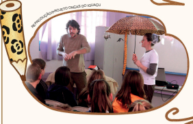 Imagem: fotografia de um homem com camisa marrom e uma mulher com camiseta branca segurando um guarda-chuva marrom com pintas com figuras de onças penduradas. Olhando para eles, crianças uniformizadas sentadas em cadeiras. À esquerda há um lápis com estampa de onça. Fim da imagem.