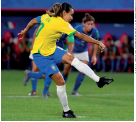 Imagem: Fotografia. Marta, mulher com cabelos presos, usando uniforme amarelo e azul. Ela está em pé com o corpo encurvado e a perna direita estendida em um chute. Atrás, outras jogadoras em um campo de futebol.  Fim da imagem.