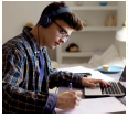 Imagem: Fotografia. Um menino com cabelos lisos, usando um fone de ouvido e casaco. Ele está sentado, com as mãos sobre a mesa. Sobre a mesa, um computador e um caderno.  Fim da imagem.