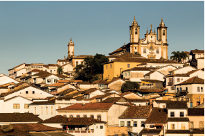 Imagem: Fotografia em tons de sépia. Cidades composta por casas sobrepostas no fundo e no topo, uma igreja com duas torres.  Fim da imagem.