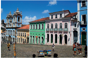 Imagem: Fotografia. Rua de paralelepípedo com casas de dois andares coloridas com muitas portas e janelas.  Fim da imagem.