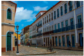 Imagem: Fotografia. Rua com prédio de três andares com muitas portas e janelas.  Fim da imagem.