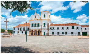 Imagem: Fotografia. Prédio branco com detalhes dourados, com muitas portas e janelas. No centro no topo, uma cruz. À esquerda, no pátio, uma escultura de uma cruz.  Fim da imagem.