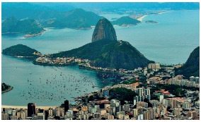 Imagem: Fotografia. Vista aérea de uma montanha e um morro cercado pelo mar. À direita, uma orla com diversos prédios.  Fim da imagem.