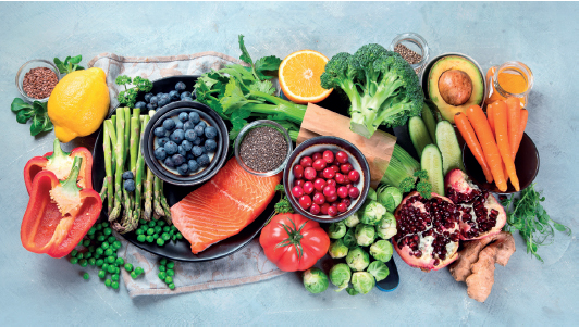 Imagem: Fotografia. Vista de diversos alimentos sobre a mesa: Pimentão, vagem, cebolinha, salmão, laranja, uva, tomate cereja, brócolis, cenoura, abacate e grãos.  Fim da imagem.