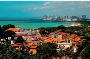 Imagem: Fotografia. Casas pequenas, ao redor, há vegetação. Atrás, o mar. No fundo, silhueta de uma cidade com grandes prédios Fim da imagem.