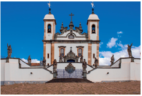 Imagem: Fotografia. Fachada de um santuário, composto por duas janelas e duas torres. Na frente, um muro com esculturas.das.  Fim da imagem.