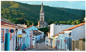 Imagem: Fotografia. Uma igreja, ao lado casas, palmeiras e vegetação. Na frente, o oceano. Atrás, montanhas com vegetação.  Fim da imagem.