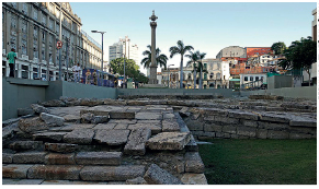 Imagem: Fotografia. Destaque de uma escada de pedras longas. Atrás, uma fonte. No fundo, silhueta de casas.  Fim da imagem.