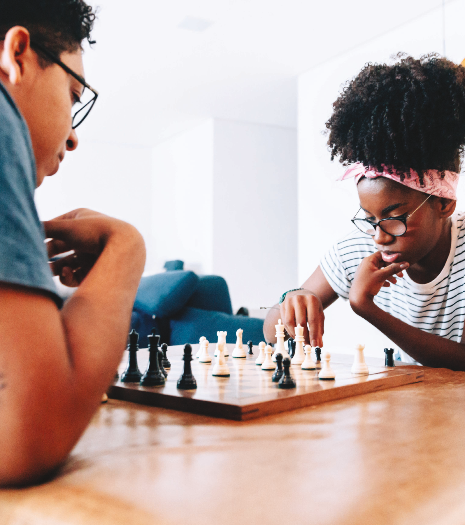 Fotografia. Duas crianças jogando xadrez. Menino negro de cabelo preto, óculos e camiseta azul. Menina negra de cabelo cacheado com uma faixa rosa na cabeça, óculos e camiseta branca listrada. Ela está com uma mão no queixo e outra em uma das peças.