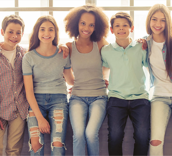 Fotografia. Cinco pessoas sentadas na frente de uma janela de vidro. No centro, menina negra de cabelo na altura dos ombros, camiseta regata cinza e calça jeans. De um lado, menino branco de cabelo castanho, camisa xadrez e calça bege e menina branca de cabelo castanho longo, camiseta azul e calça jeans. Do outro lado, menino branco de cabelo castanho, camisa verde e calça azul e menina branca de cabelo castanho longo, camiseta branca, jaqueta e calça jeans. Todos eles estão sorrindo.