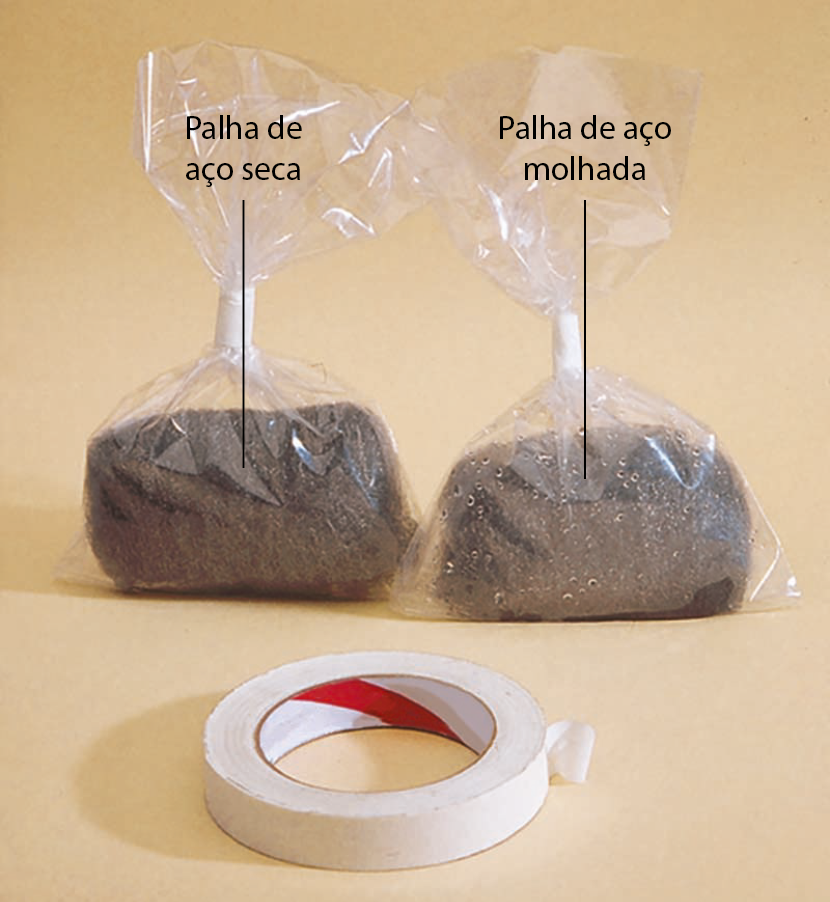 Ilustração. Dois sacos de plástico transparente fechados com um material metálico retangular esponjoso dentro. Em um deles, palha de aço seca. No outro, palha de aço molhada. Na frente, um rolo de fita adesiva branca.