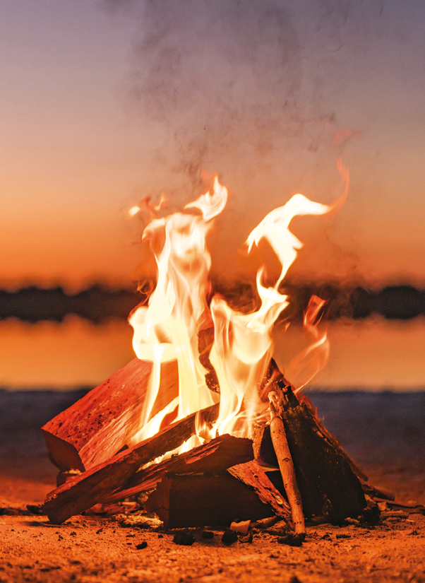 Fotografia. Uma fogueira no chão com o fogo em toras de madeira no cair da noite.