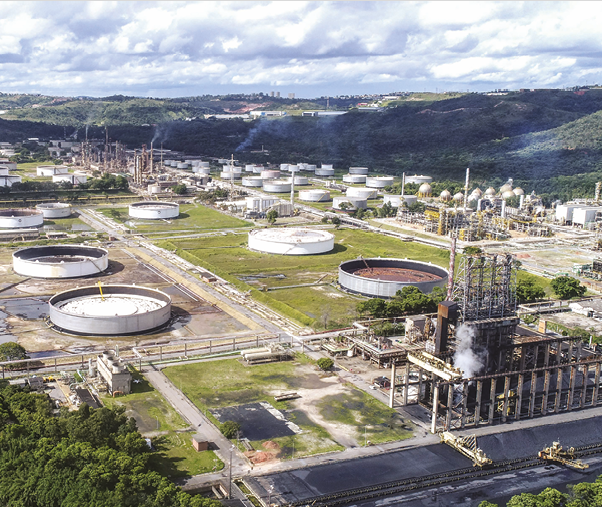 Fotografia. Um parque industrial com chaminés saindo fumaça e tanques cilíndricos espaçados.